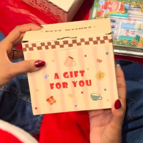 A person holding a small cream-colored gift box with red text reading 'A GIFT FOR YOU' and a checkered border, with other matching gift boxes and a colorful item in the background on a red surface.