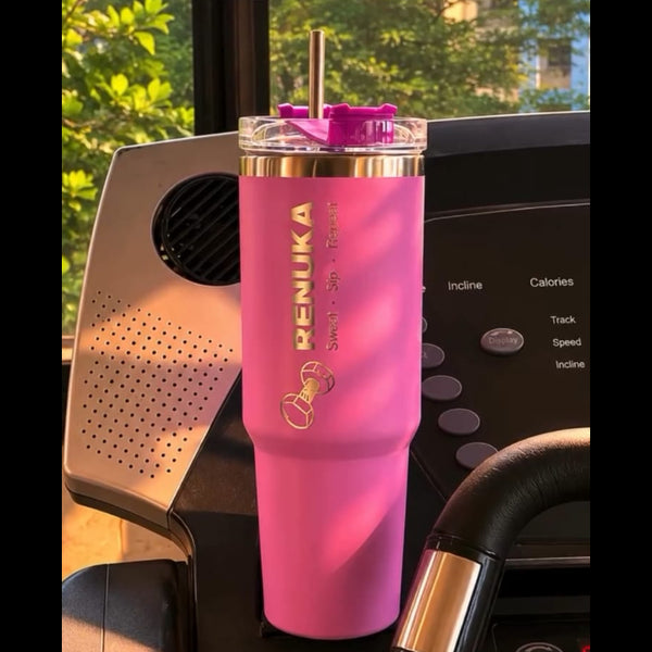 A pink insulated tumbler with a clear lid and straw, featuring gold lettering that reads 'RENUKA' and '16 oz', shown resting on a black exercise bike or treadmill in a real indoor environment with sunlight and window views in the background.