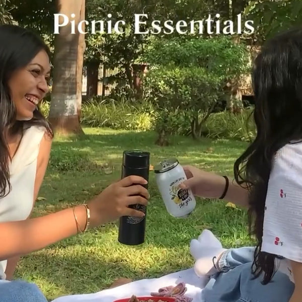 Two women sitting on a white picnic blanket in a park, clinking a black insulated tumbler and a white can. The scene has the text 'Picnic Essentials' overlaid at the top, with grass, trees, and picnic items like bread, a red bowl, and a cup visible.