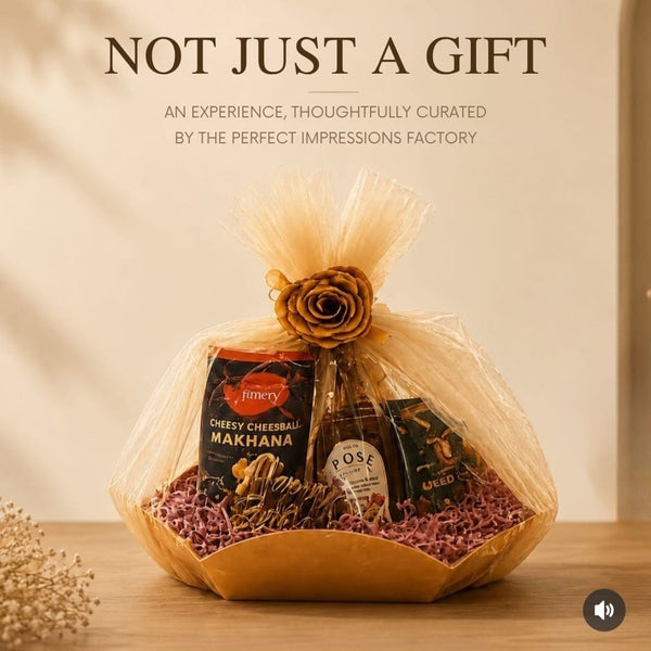 A curated gift basket wrapped in sheer beige tulle with a decorative rose, displayed on a wooden surface against a warm beige background. Visible items include a black box of Darjeeling Makhana, a small bottle of Rose Hip powder, and other packaged snacks.