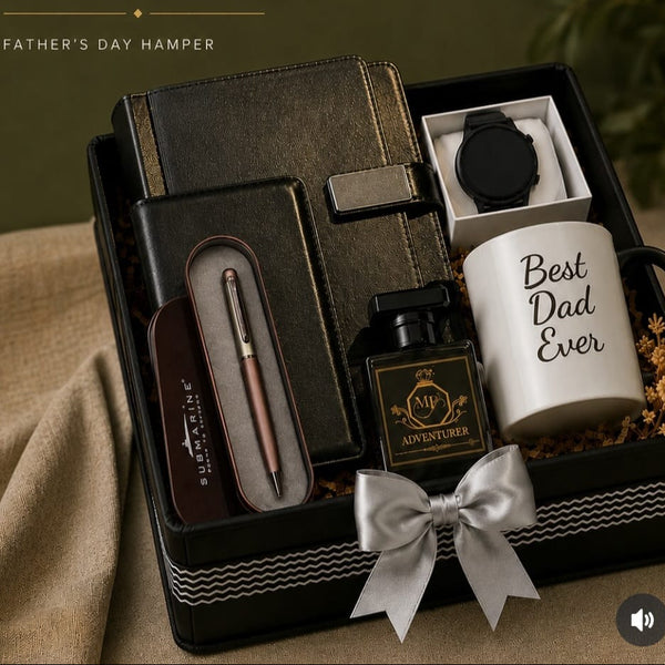 A curated Father's Day gift set displayed in a black presentation box with a white bow. The set includes a dark leather notebook, a pen in a case, a small black bottle labeled 'ADVENTURE', a white mug reading 'Best Dad Ever', and a black round accessory in a white insert.